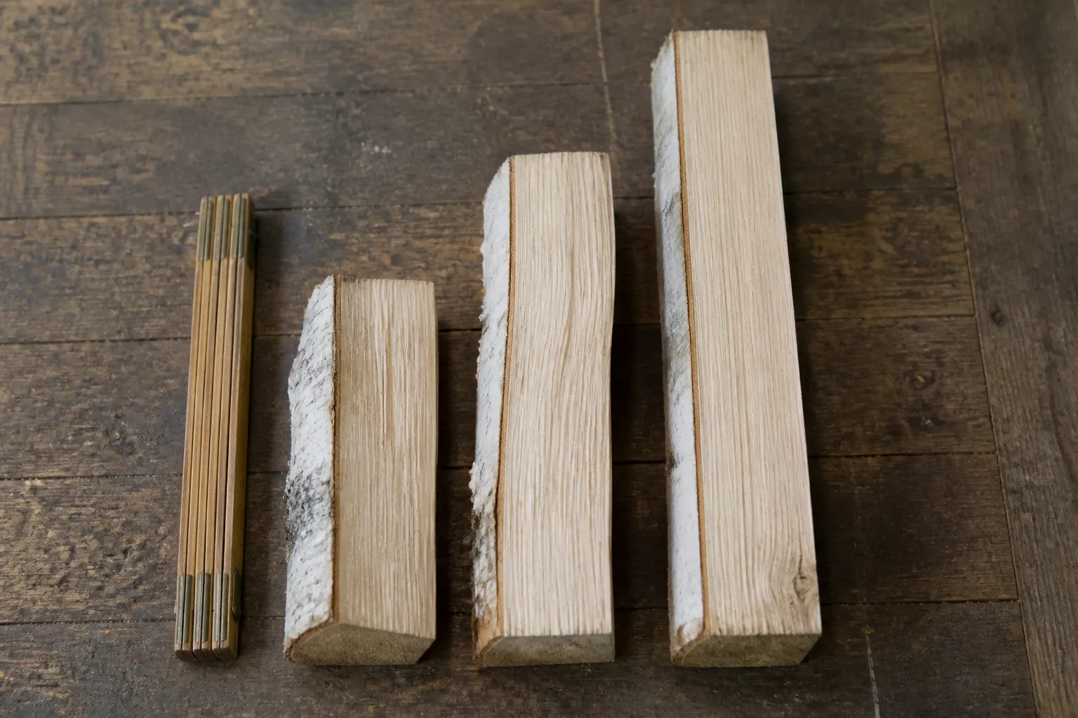 Drei Holzscheite unterschiedlicher Länge (25 cm, 33 cm und 50 cm) mit einem Zollstock daneben, auf einem Holzboden